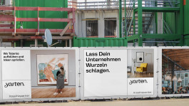 Bauzaunbanner an einer Baustelle, das das Kreativquartier Potsdam bewirbt. Zu sehen sind verschiedene Motive: eine Kunstgalerie, eine moderne Inneneinrichtung und ein Café, kombiniert mit den Slogans „Wo Talente aufblühen und Ideen sprießen.“ und „Lass Dein Unternehmen Wurzeln schlagen.“