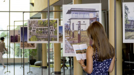 Eine Frau mit langen Haaren und gepunktetem Kleid steht in einer Ausstellung und hält eine Broschüre in der Hand. Sie betrachtet ein großformatiges Foto eines historischen Ortes, das an einer Metallkonstruktion hängt. Weitere Bilder und eine andere Besucherin sind im Hintergrund zu sehen.
