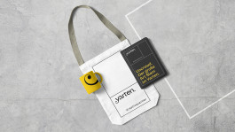 Flach liegende Stofftasche auf grauem Betonhintergrund mit schwarzem „yarten. Kreativquartier“-Aufdruck. Darauf liegen ein schwarzes Notizbuch mit gelber Typografie und ein gelbes Spiralarmband mit einem schwarzen grafischen Element.