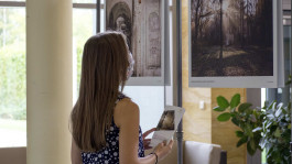 Eine Frau mit langen Haaren und gepunktetem Kleid steht in einer Ausstellung und betrachtet zwei großformatige Fotografien historischer Orte. Sie trägt eine Maske und hält eine Broschüre mit dem Titel der Ausstellung »Früher Sperrzone, heute Ausflugsziel«.