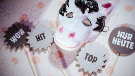 Flache Requisiten für ein Video-Shopping-Format, darunter eine weiße Pferdemaske und Pappschilder mit den Worten „WOW“, „HIT“, „TOP“ und „NUR HEUTE“.